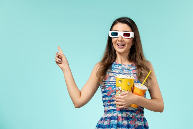 Vista frontal mujer joven sosteniendo bebida de palomitas de maíz en gafas de sol d sobre la superficie azul