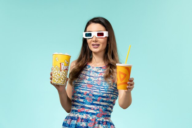 Vista frontal mujer joven sosteniendo bebida de palomitas de maíz en gafas de sol d sobre la superficie azul