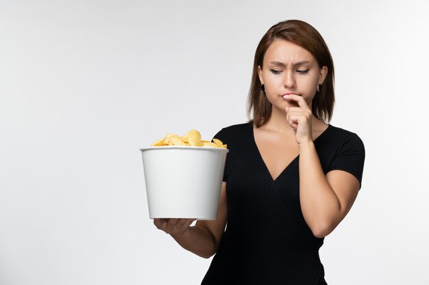 Foto gratuita vista frontal mujer joven sosteniendo la canasta con papas fritas y comiendo en la superficie blanca clara