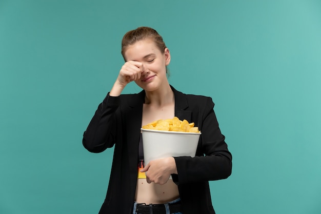 Vista frontal mujer joven sosteniendo y comiendo patatas fritas viendo la película llorando en la superficie azul