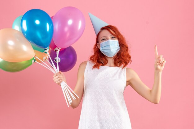 Vista frontal de la mujer joven sosteniendo globos de colores en máscara estéril en la pared rosa