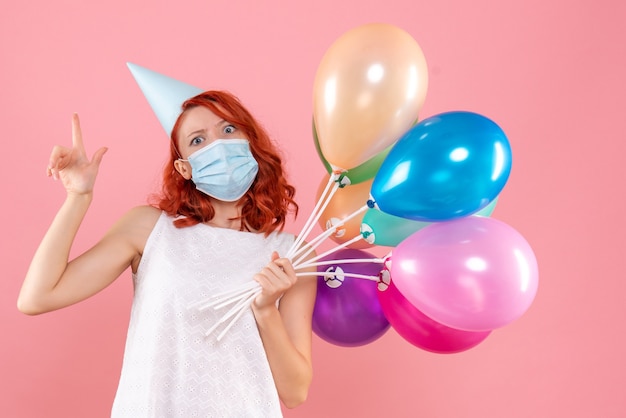 Vista frontal de la mujer joven sosteniendo globos de colores en máscara estéril en la pared rosa