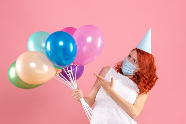 Vista frontal de la mujer joven sosteniendo globos de colores en máscara estéril en la pared rosa