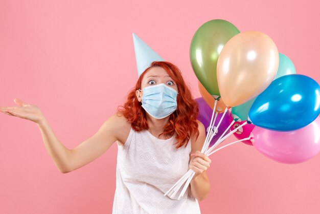 Vista frontal de la mujer joven sosteniendo globos de colores en máscara estéril en la pared rosa