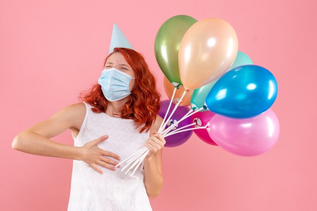 Vista frontal de la mujer joven sosteniendo globos de colores en máscara estéril en la pared rosa