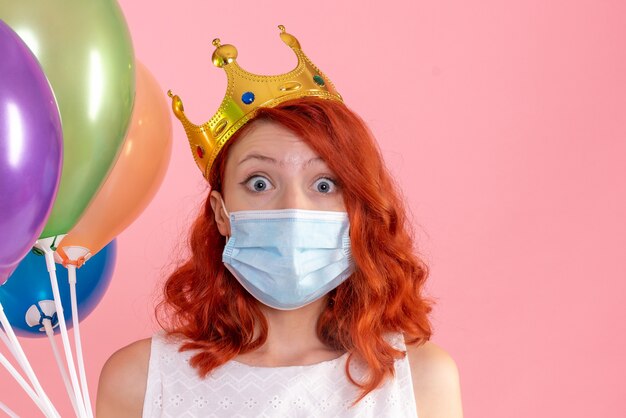 Vista frontal de la mujer joven sosteniendo globos de colores en máscara estéril en la pared rosa