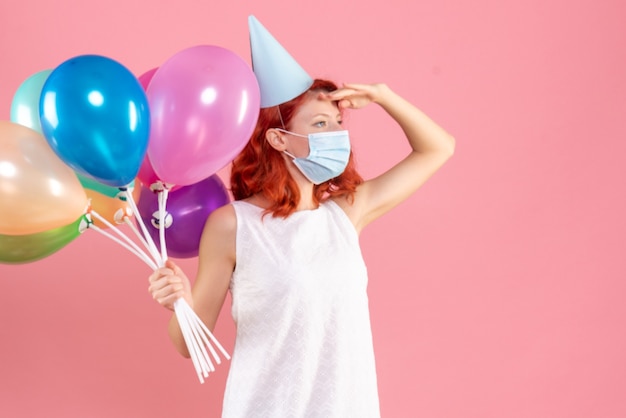 Vista frontal de la mujer joven sosteniendo globos de colores en máscara estéril en la pared rosa