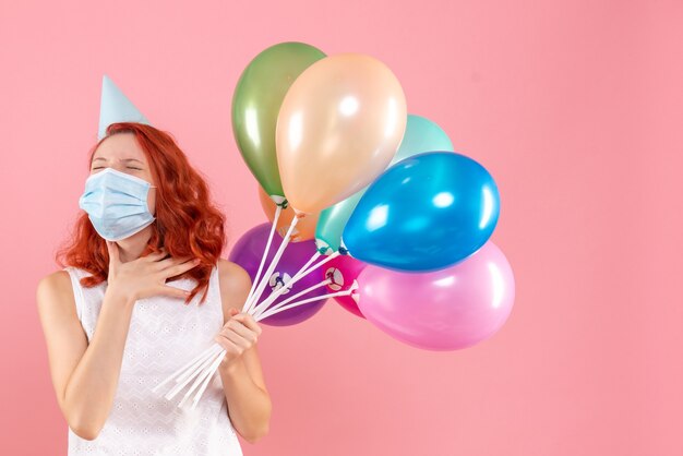 Vista frontal de la mujer joven sosteniendo globos de colores en máscara estéril en la pared rosa