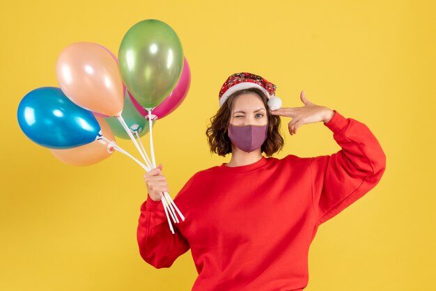 Vista frontal mujer joven sosteniendo globos en máscara estéril en piso amarillo celebración de fiesta de emoción de color de año nuevo mujer