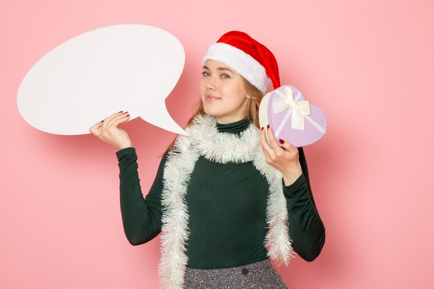 Vista frontal mujer joven sosteniendo un gran cartel blanco y presente en la pared rosa modelo vacaciones navidad año nuevo color emoción