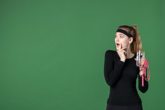 Vista frontal de la mujer joven sosteniendo la medida de la cintura y un vaso de agua sobre fondo verde, color, salud, cuerpo, entrenamiento, mujer, atleta, negro