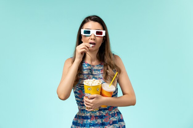 Vista frontal mujer joven sosteniendo paquete de palomitas de maíz y bebida en gafas de sol d sobre la superficie azul