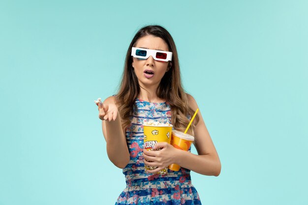 Vista frontal mujer joven sosteniendo paquete de palomitas de maíz y bebida en gafas de sol d sobre la superficie azul