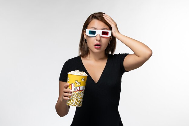 Vista frontal mujer joven sosteniendo el paquete de palomitas de maíz en gafas de sol d y viendo una película en una superficie blanca