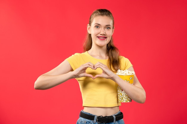 Vista frontal mujer joven sosteniendo el paquete de palomitas de maíz y mostrando el signo del corazón en la pared de color rojo claro película cine cine