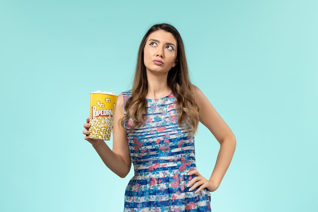 Vista frontal mujer joven sosteniendo el paquete de palomitas de maíz y pensando en la superficie azul