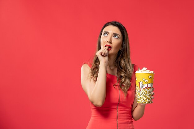 Vista frontal mujer joven sosteniendo el paquete de palomitas de maíz pensando en la superficie roja