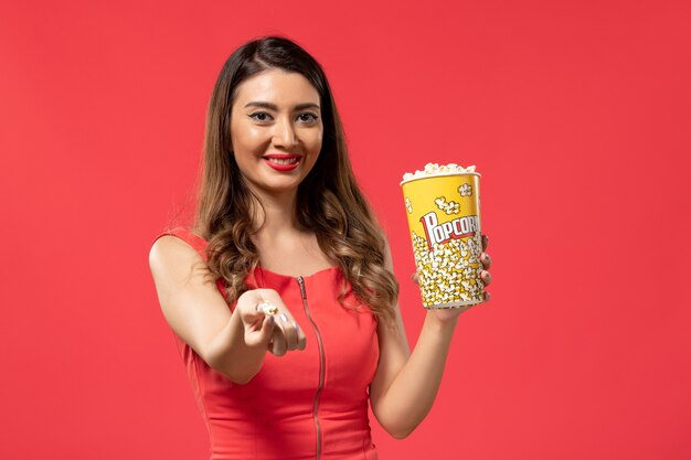 Vista frontal mujer joven sosteniendo el paquete de palomitas de maíz y sonriendo sobre superficie roja