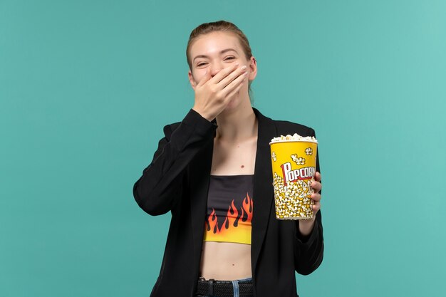 Vista frontal mujer joven sosteniendo el paquete de palomitas de maíz viendo la película y riendo en la superficie azul