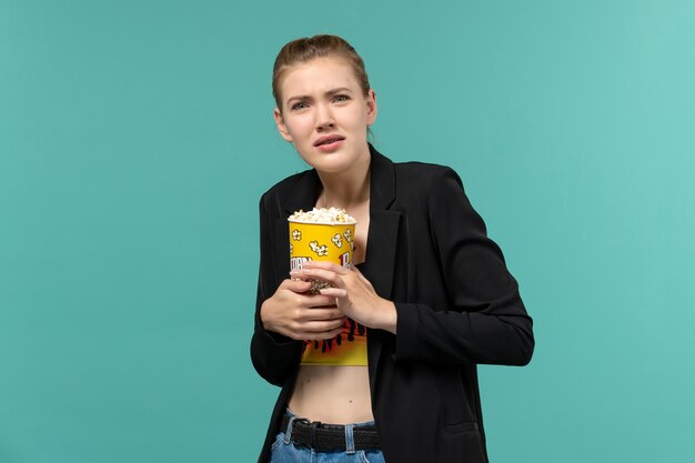 Vista frontal mujer joven sosteniendo el paquete de palomitas de maíz viendo la película en la superficie azul claro