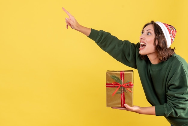 Vista frontal de la mujer joven sosteniendo un pequeño regalo de Navidad en la pared amarilla