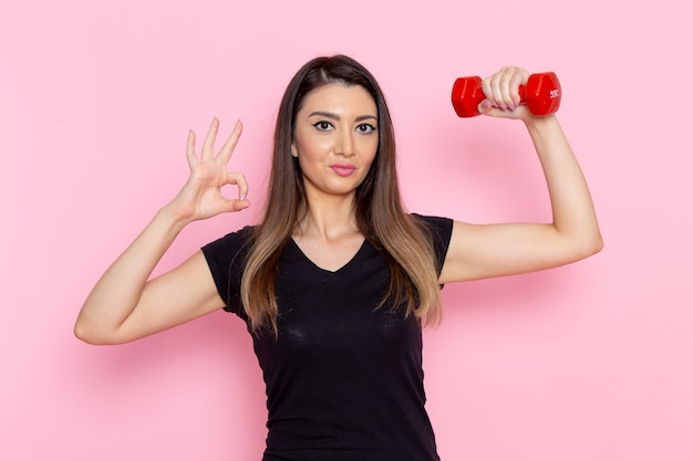 Vista frontal mujer joven sosteniendo pesas rojas en la pared rosa claro atleta deporte ejercicio salud entrenamiento