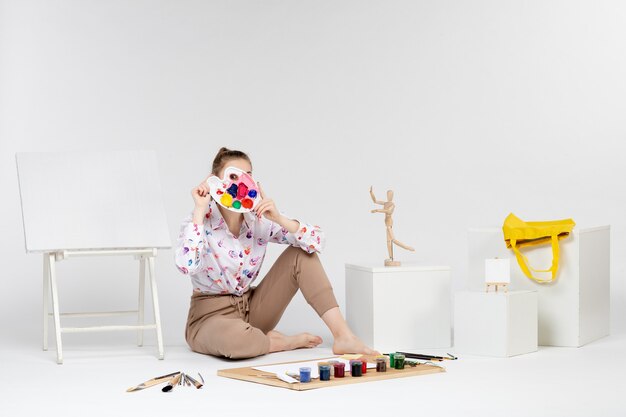 Vista frontal mujer joven sosteniendo pinturas para dibujar sobre fondo blanco.