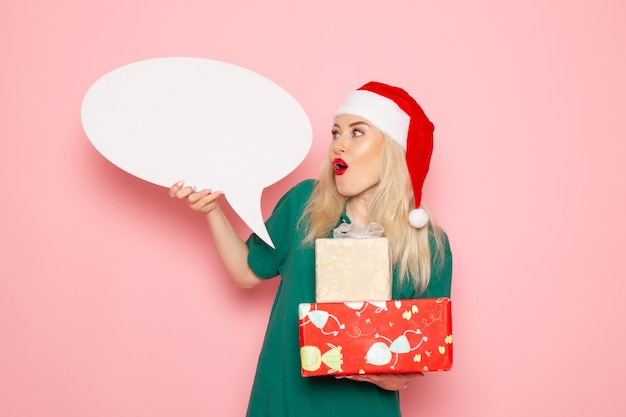 Vista frontal mujer joven sosteniendo regalos de Navidad y cartel blanco en la pared rosa mujer regalo nieve foto en color vacaciones de año nuevo