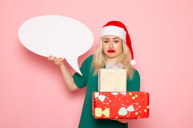 Vista frontal mujer joven sosteniendo regalos de Navidad y letrero blanco en la pared rosa mujer regalo nieve color foto año nuevo