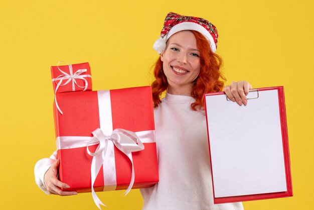 Vista frontal de la mujer joven sosteniendo regalos de Navidad y nota de archivo en la pared amarilla