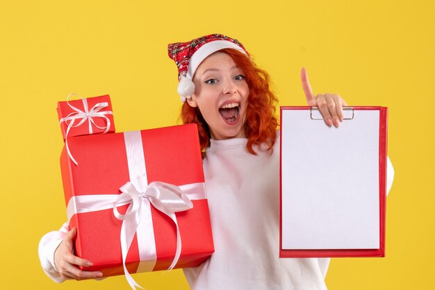 Vista frontal de la mujer joven sosteniendo regalos de Navidad y nota de archivo en la pared amarilla