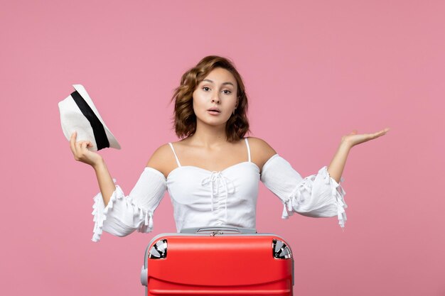 Vista frontal de la mujer joven sosteniendo el sombrero y preparándose para el viaje con bolsa roja en la pared rosa