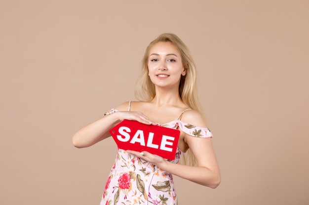 Vista frontal de la mujer joven sosteniendo el tablero de venta rojo en la pared marrón