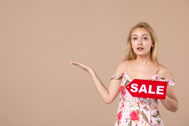 Vista frontal de la mujer joven sosteniendo el tablero de venta rojo en la pared marrón