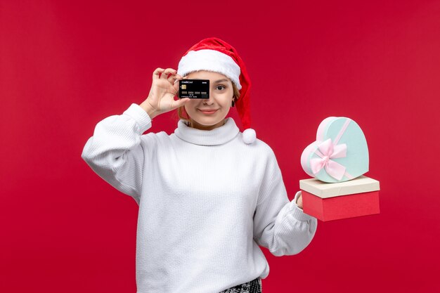 Vista frontal mujer joven con tarjeta bancaria y regalos sobre fondo rojo.