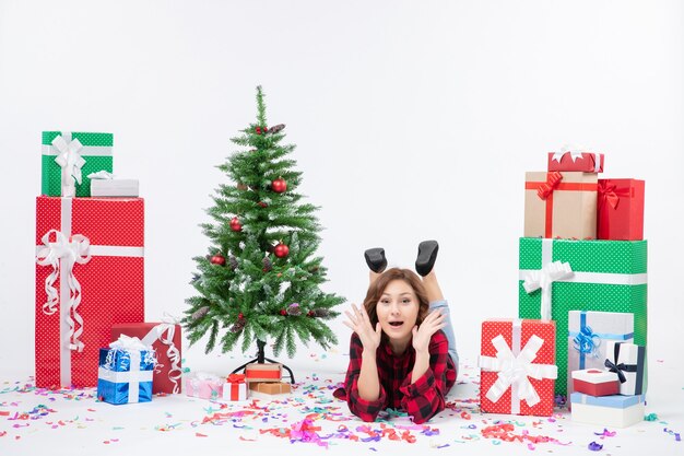 Vista frontal mujer joven tendido alrededor de regalos de navidad y árbol de vacaciones en el escritorio blanco regalo de año nuevo de navidad color nieve