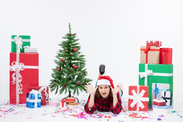 Vista frontal de la mujer joven tendido alrededor de regalos de Navidad y arbolito de vacaciones en la pared blanca