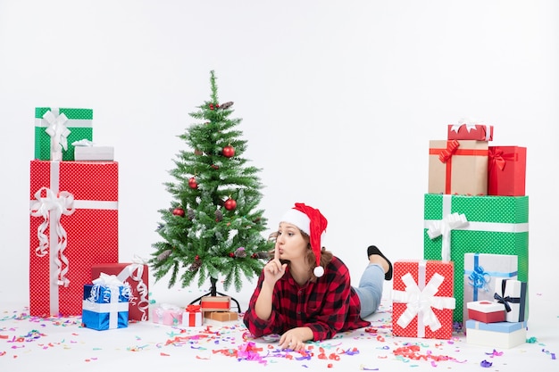 Vista frontal de la mujer joven tendido alrededor de regalos de Navidad y arbolito de vacaciones en la pared blanca