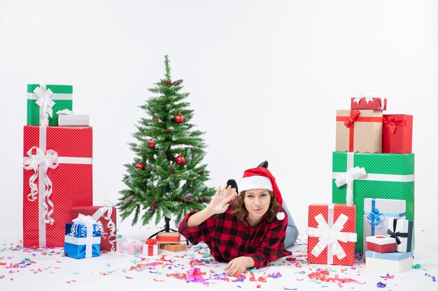 Vista frontal mujer joven tendido alrededor de regalos de navidad y arbolito de vacaciones sobre fondo blanco año nuevo mujer fría emoción de navidad nieve