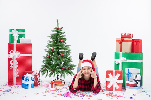 Vista frontal mujer joven tendido alrededor de regalos de Navidad y arbolito de vacaciones sobre fondo blanco mujer fría Navidad año nuevo nieve