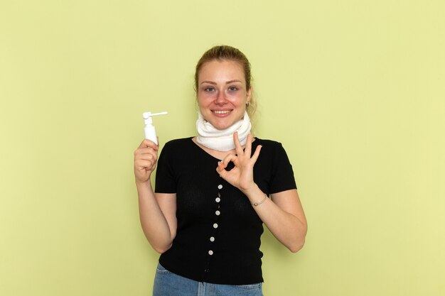 Vista frontal mujer joven con una toalla blanca alrededor de su garganta sintiéndose muy enferma y enferma sosteniendo spray para la garganta en el escritorio verde claro enfermedad salud femenina