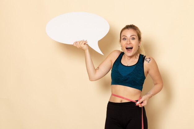 Vista frontal mujer joven en traje deportivo con cartel blanco y midiendo su cuerpo en la pared beige claro, salud corporal, deporte, ejercicios de belleza en forma