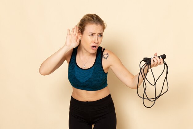 Foto gratuita vista frontal mujer joven en traje deportivo sosteniendo cuerdas para saltar y escuchando en la pared blanca ejercicios de belleza de cuerpo de entrenamiento de salud