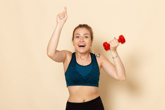 Foto gratuita vista frontal mujer joven en traje deportivo sosteniendo pesas rojas en la pared blanca luz deporte cuerpo salud belleza entrenamiento