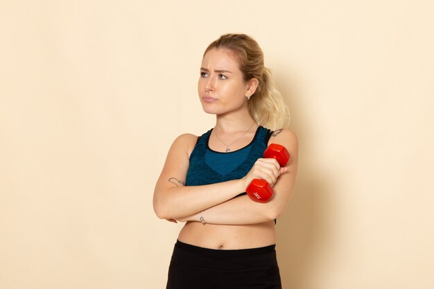 Vista frontal mujer joven en traje deportivo sosteniendo pesas rojas en la pared blanca salud deporte cuerpo belleza entrenamiento