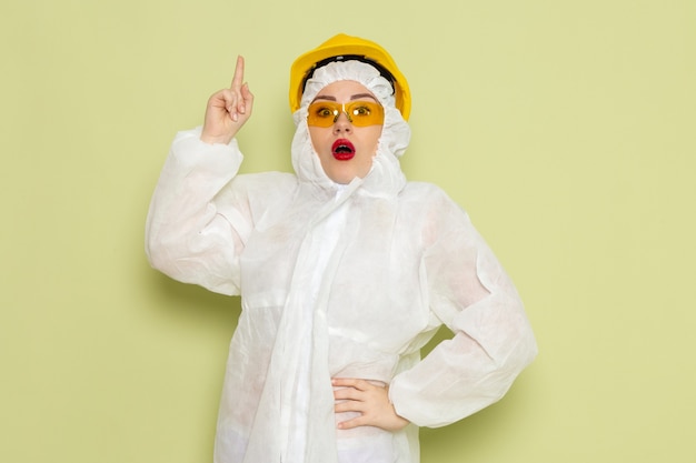 Vista frontal mujer joven en traje especial blanco y casco amarillo posando con el dedo levantado en el trabajo del espacio verde