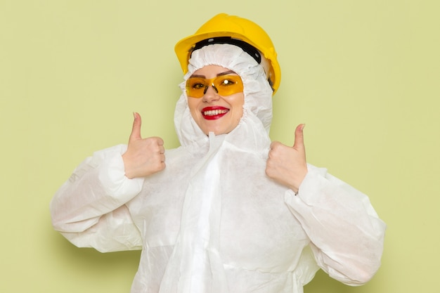 Foto gratuita vista frontal mujer joven en traje especial blanco y casco amarillo sonriendo y mostrando como signo en el trabajo del espacio verde