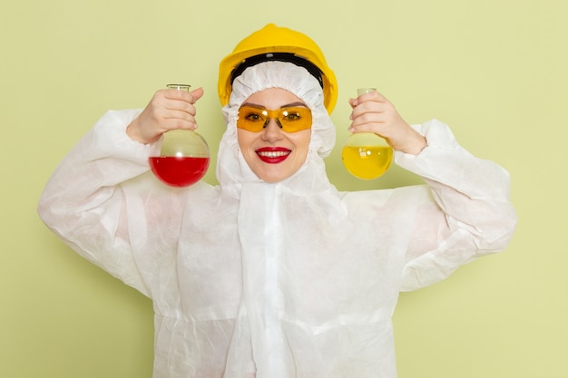 Vista frontal mujer joven en traje especial blanco y casco amarillo sosteniendo frascos con soluciones en el espacio verde