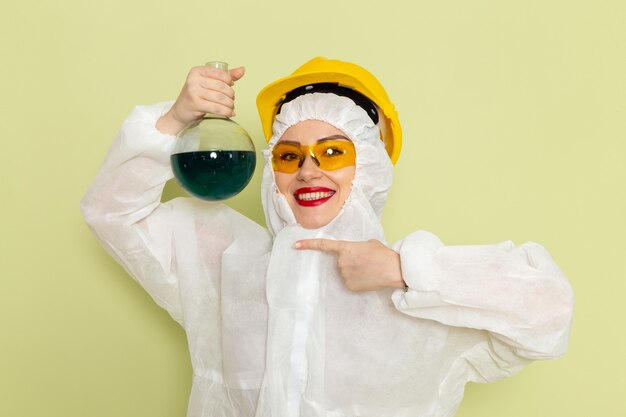 Vista frontal mujer joven en traje especial blanco y casco amarillo sosteniendo el matraz y sonriendo en el espacio verde