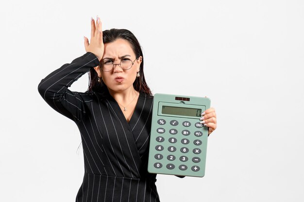 Vista frontal mujer joven en traje oscuro estricto con gran calculadora sobre fondo blanco trabajo belleza mujer moda oficina de negocios